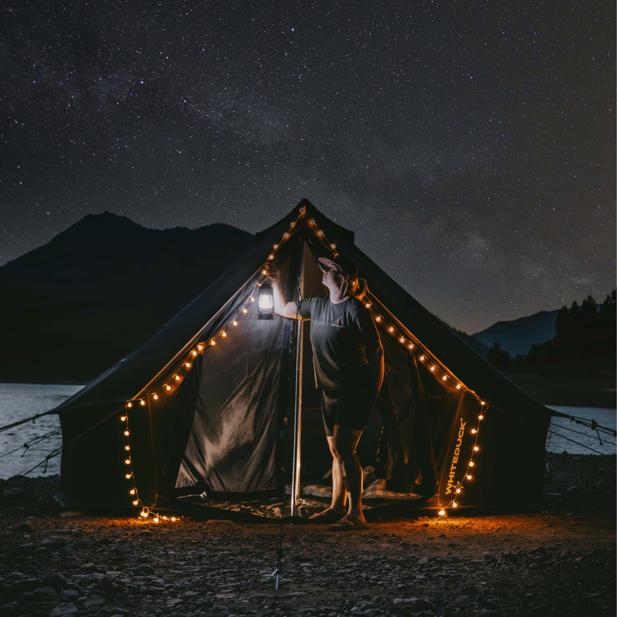White Duck Outdoors 13' Regatta Bell Tent under starry night sky with fairy lights and a woman holding a lantern.