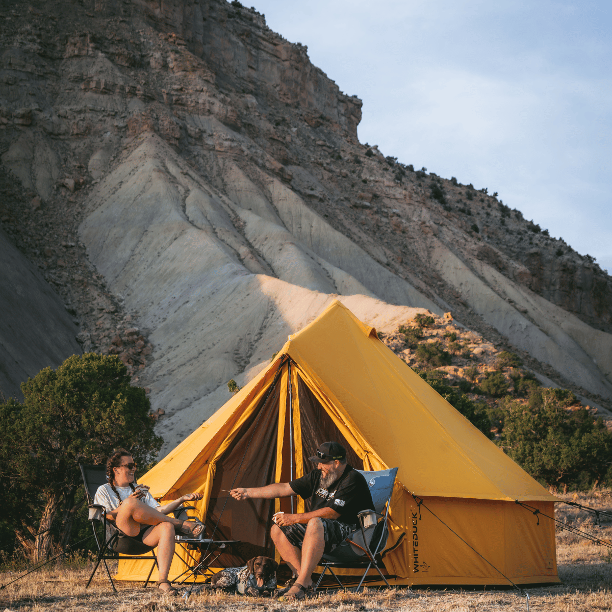 White Duck Outdoors 16-foot Regatta Bell Tent set up outdoors with a couple, featuring a Tuscan Sun backdrop and mountain scenery.