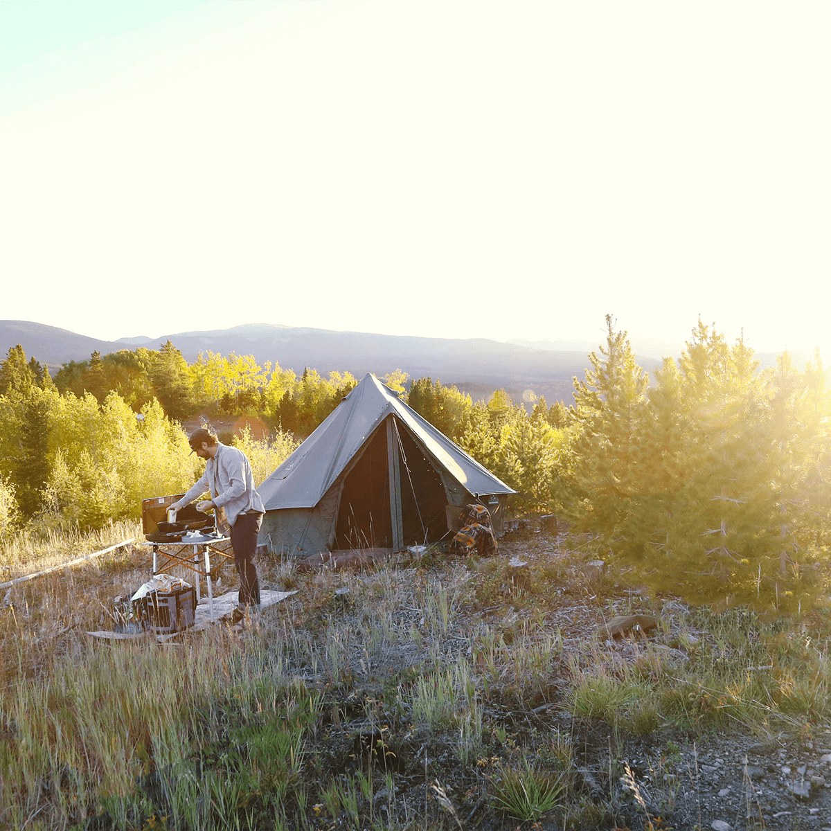 White Duck Outdoors 13' Regatta Bell Tent in Boulder Grey with a man cooking outside, showcasing camping and outdoor living.