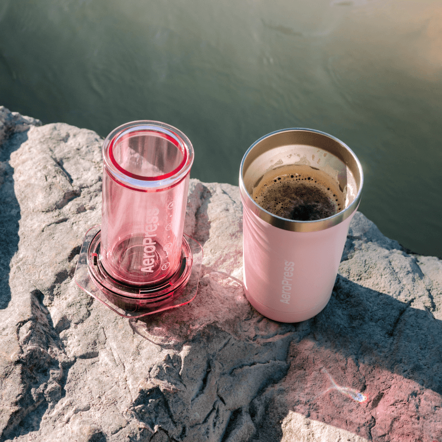 Two travel mugs, one pink and one red with 'Aeropress' branding, on a rock with a scenic background.