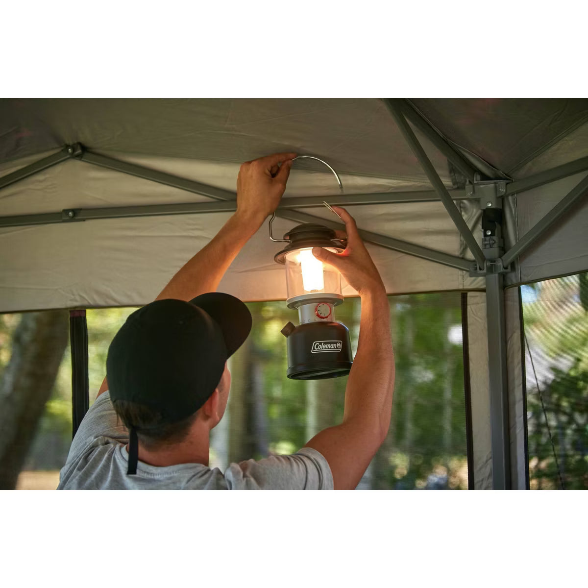 Person hanging a Coleman lantern inside a tent with trees in the background