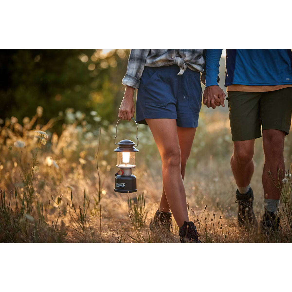 A couple walking hand in hand through a grassy field at sunset, with the woman holding a lit Camping Light lantern. The woman wears blue shorts and a plaid shirt, and the man wears blue and olive green shorts.