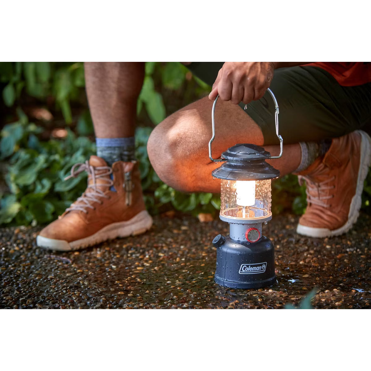 Person holding a Coleman lantern outdoors on a natural ground surface.