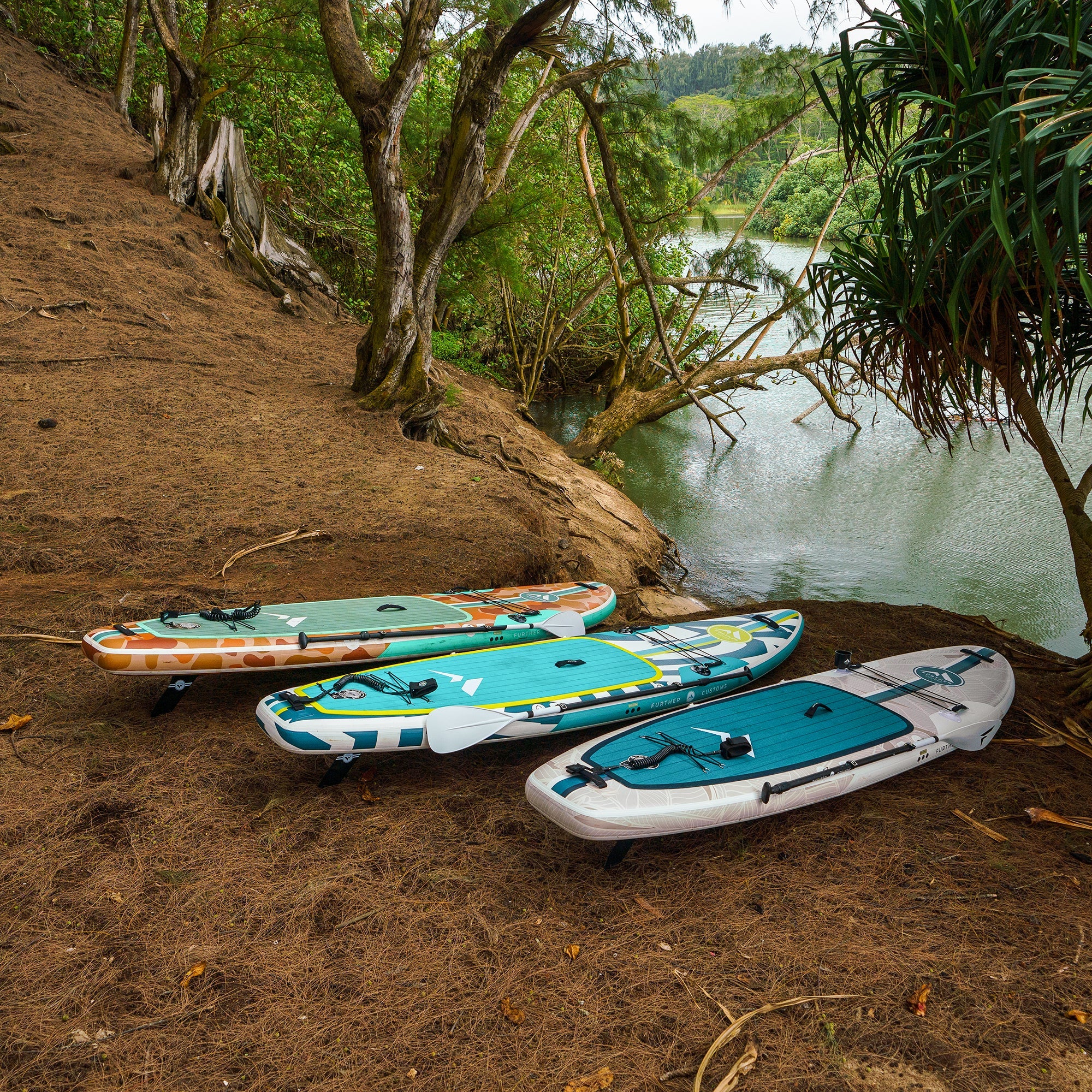 Turquoise 11'3 Podium paddleboard by Further Customs resting on a sandy beach with lush greenery in the background.