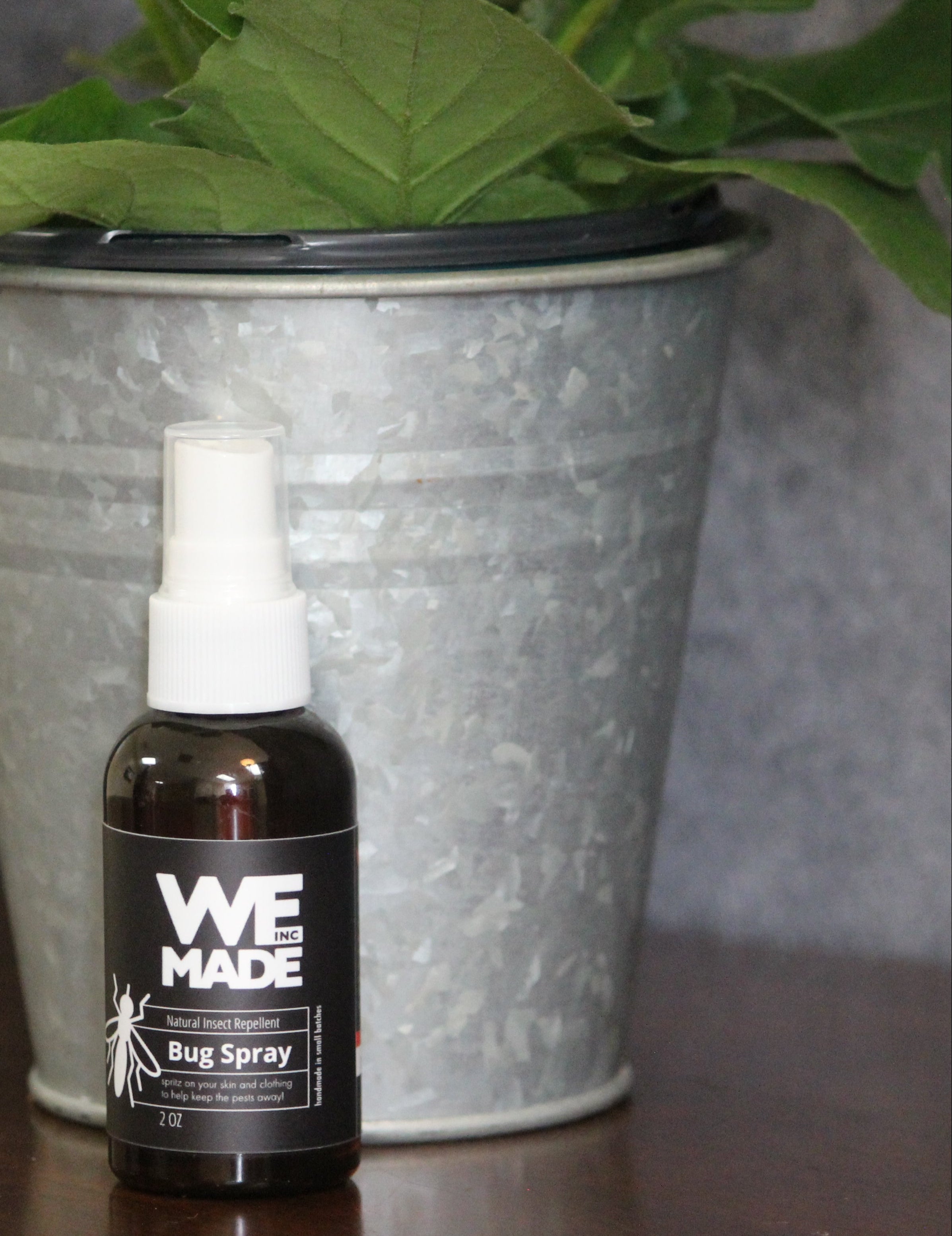 Bug spray bottle labeled 'WE MADE' on a wooden surface with a plant in the background.