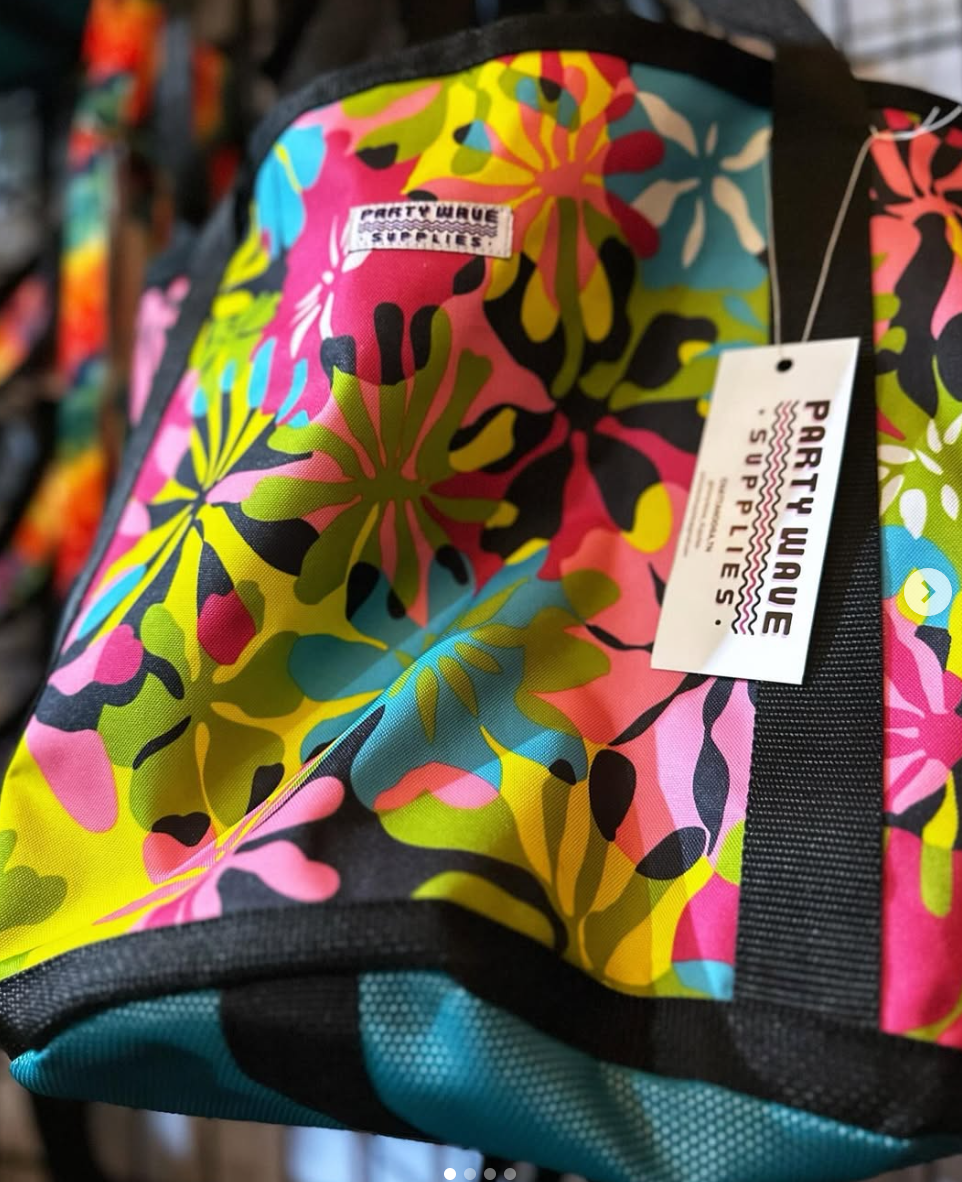 A close-up of a colorful tote bag with a bold floral pattern in pink, yellow, green, blue, and black. A white hangtag reading 'PARTY WAVE SUPPLETS' is attached, and the bag is photographed in a retail setting.