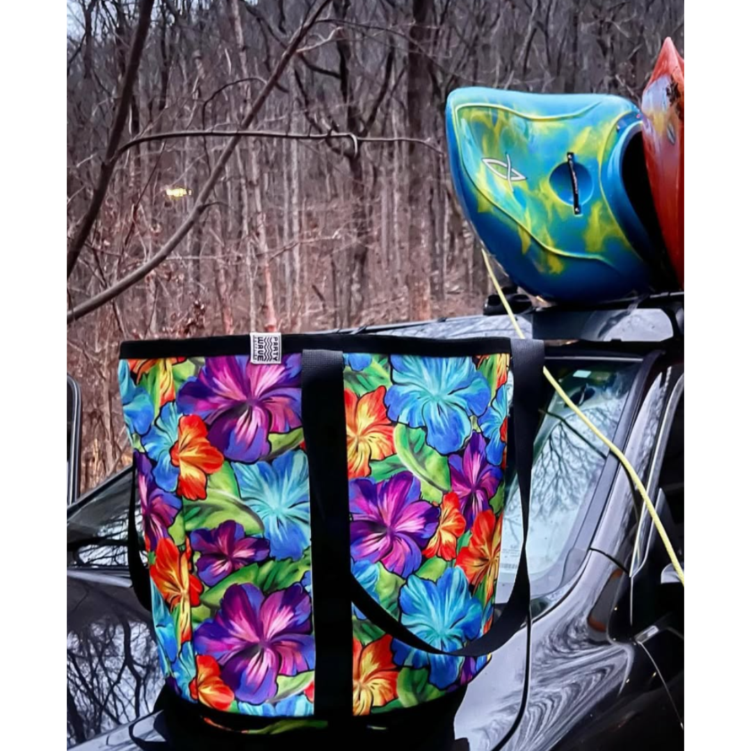 A large tote bag with a colorful tropical floral print is strapped to the roof of a black vehicle, with a blue and yellow kayak resting on top. The background shows a wooded area with bare trees.