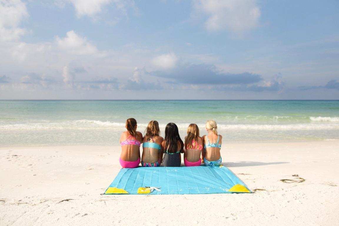 Yellow Pike Trail pocket blanket spread on the beach with four girls sitting on it, showcasing a compact outdoor blanket for camping or picnics.