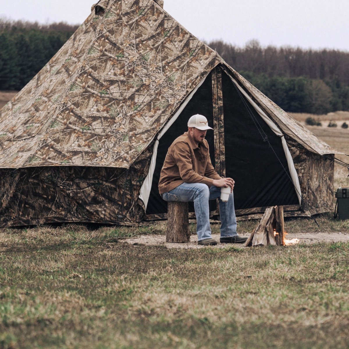 White Duck Outdoors 20-foot Regatta Bell Tent set outdoors with a man sitting on a log, featuring Realtree Edge camouflage fabric.