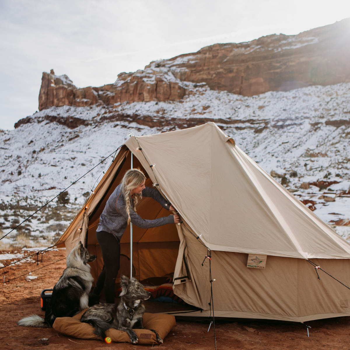Sandstone beige 13-foot Regatta Bell Tent by White Duck Outdoors set against snowy mountains with a girl and her dogs outside.