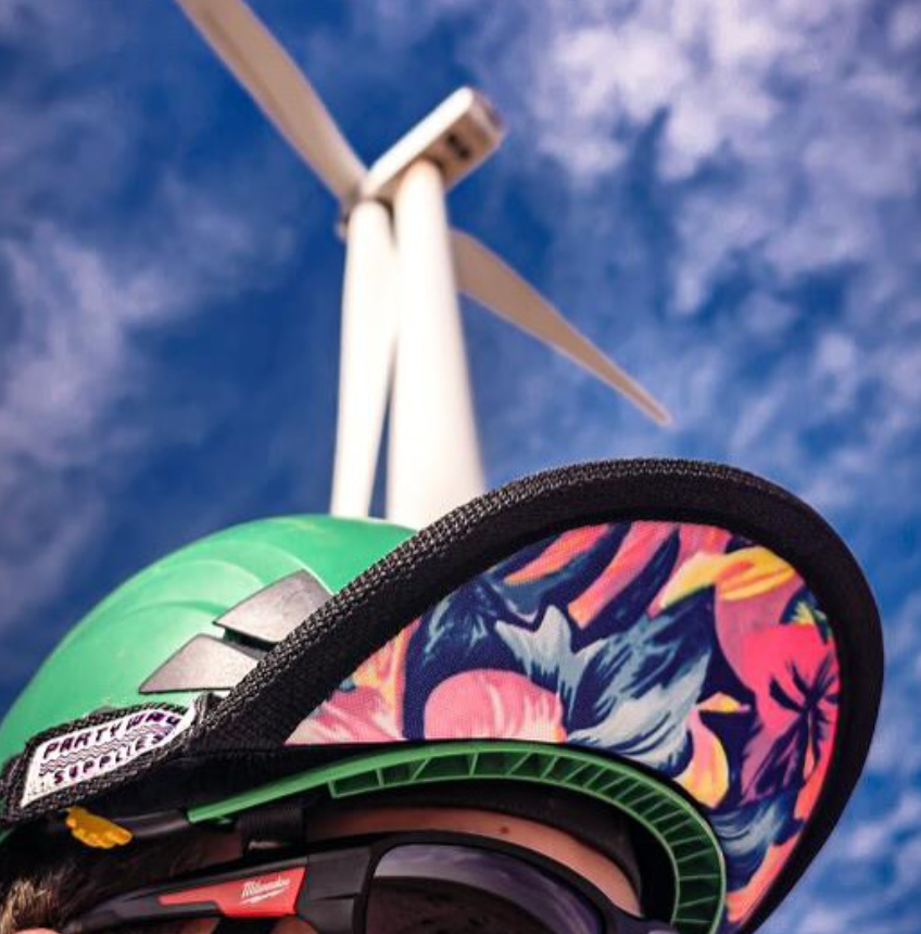 A close-up of a green and black helmet with a colorful tropical pattern visor, photographed outdoors against a blue sky and a wind turbine.
