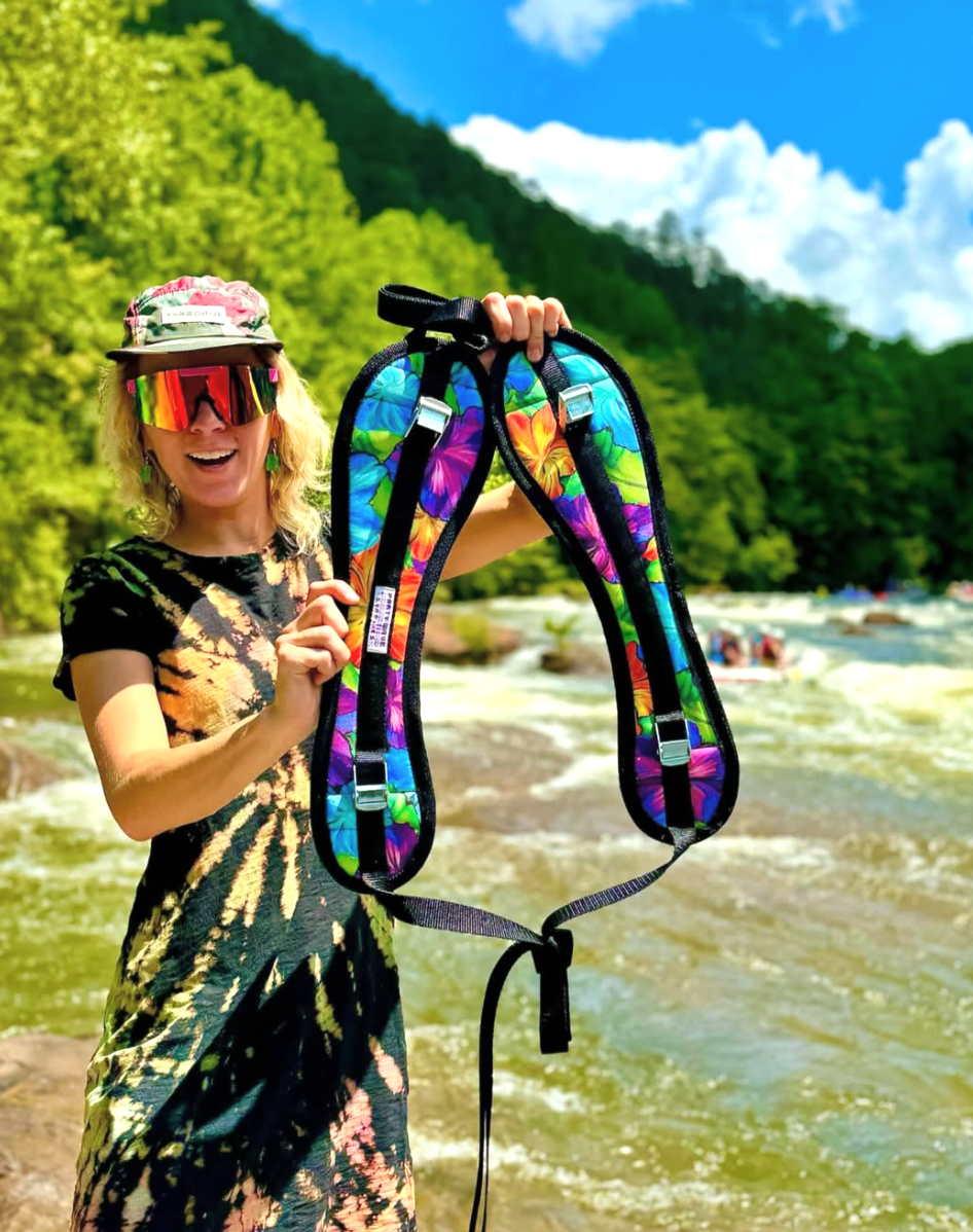 A woman standing on a riverbank in a natural outdoor setting holds up a colorful, tie-dye padded harness vest with black trim and straps. The background features rushing water, green hills, and a blue sky with clouds.