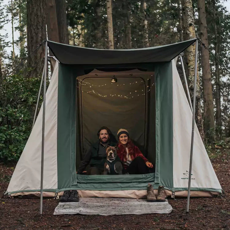 Couple and their dog relaxing outside a 7x9 Prota Canvas Cabin Tent by White Duck Outdoors in a natural outdoor setting.