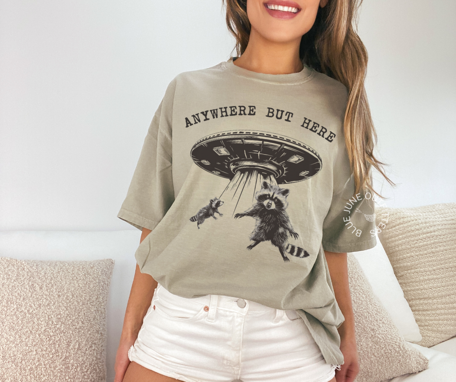 Comfort Colors khaki T-shirt featuring a funny raccoon graphic from Blue June Outfitters' Anywhere But Here UFO collection.