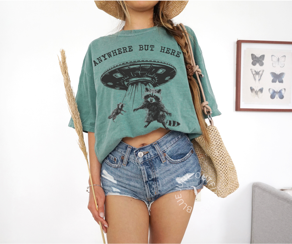 Comfort Colors light green T-shirt featuring a humorous raccoon design from Blue June Outfitters' Anywhere But Here UFO collection.