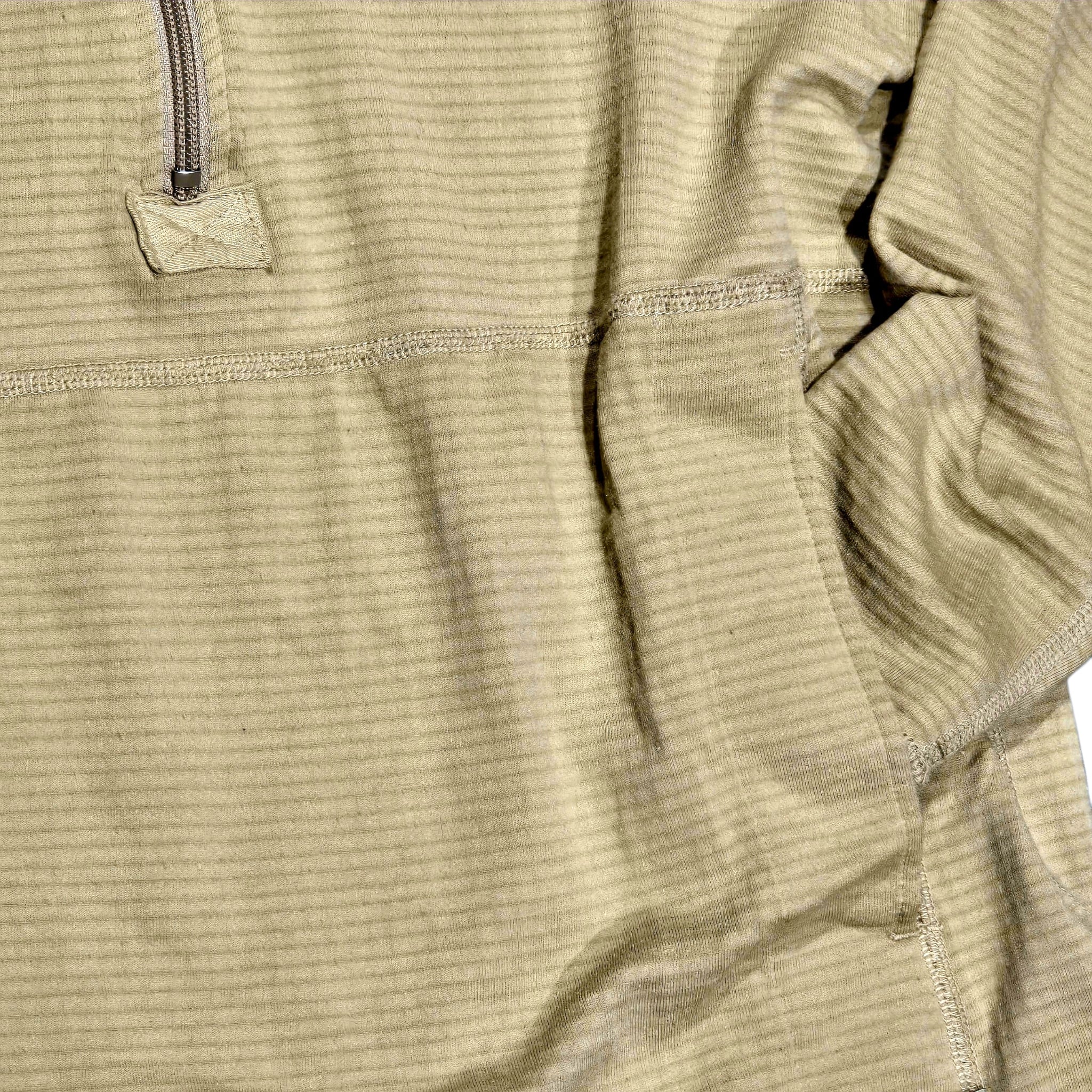 Close-up of a ribbed knit top in a muted khaki-beige color, showing the texture of the fabric, a zipper tab, and seam details.