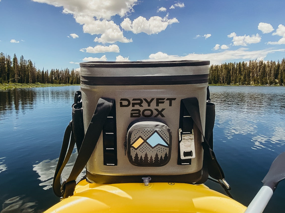 SolVida DRYFT BOX floating cooler with Bluetooth speaker, waterproof water sports accessory, shown on water with kayak in background.