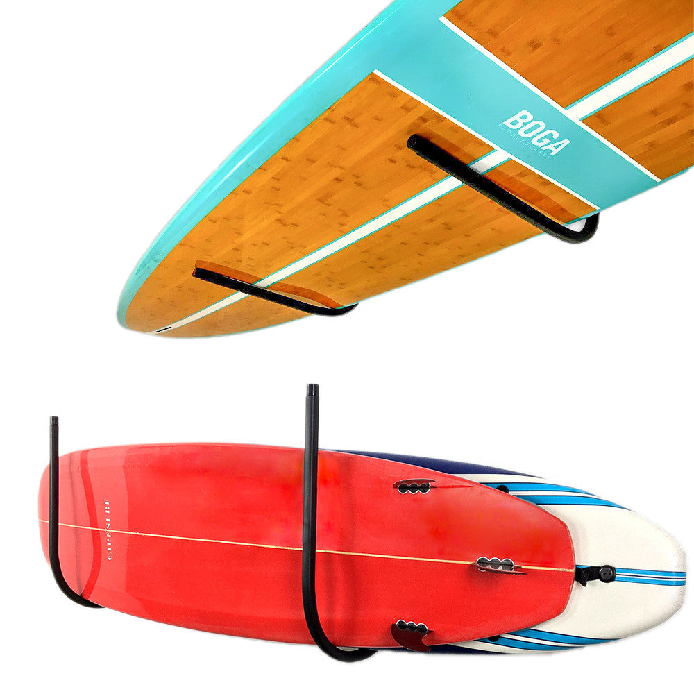 Two stand-up paddleboards shown against a white background, each with a black wall-mounted rack holding the board. The top board is turquoise and wood-toned with the brand name BOGA visible near the nose.